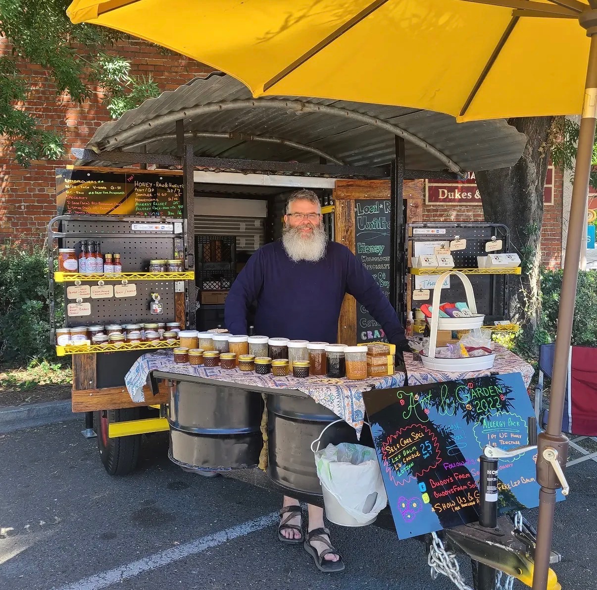 Gerald smiling behind the Buddy's Farm honey stand under a yellow umbrella